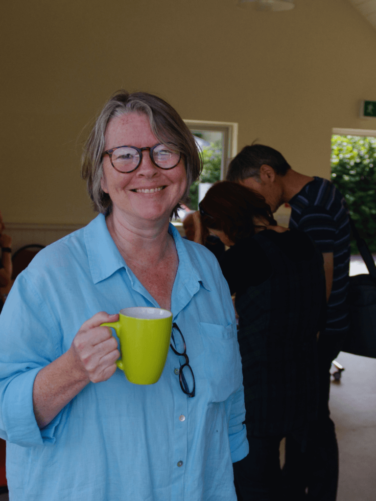 A lady with a cup of tea smiles at the camera in a community building.