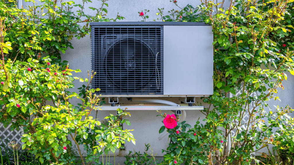 An image of a heat pump on the side of a house surrounded by vegetation.